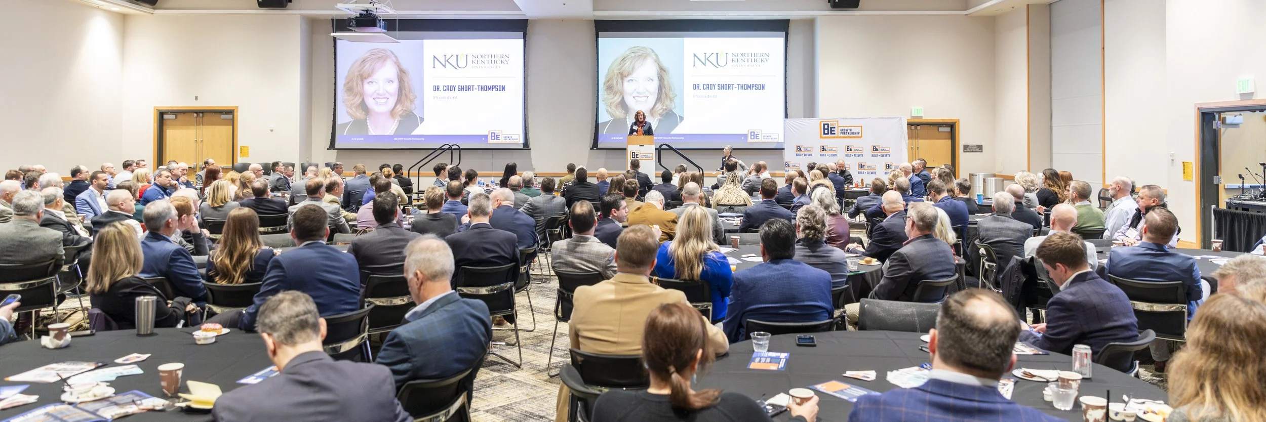 A large conference room filled with people sitting at round tables, listening to a speaker on stage. The speaker is standing at a podium with two large screens behind her displaying her photo and name, Dr. Cady Short-Thompson, and the logo of Norther