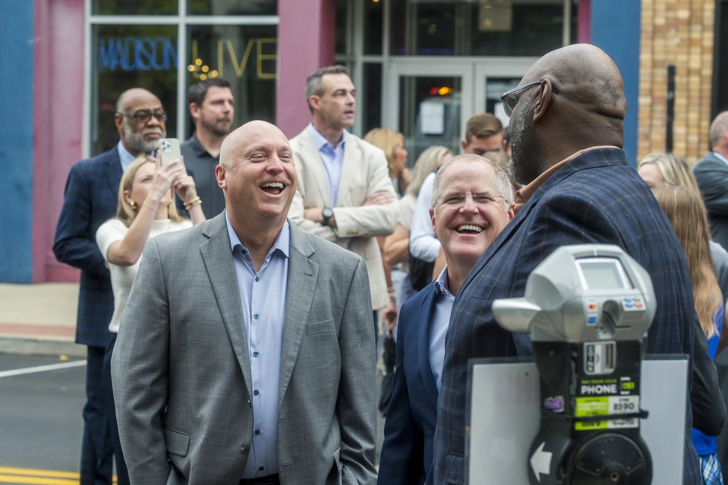 A group of people, mostly men, dressed in business attire, are smiling and laughing outdoors during a social event in front of a building with a sign that says 'MADISON LIVE'.