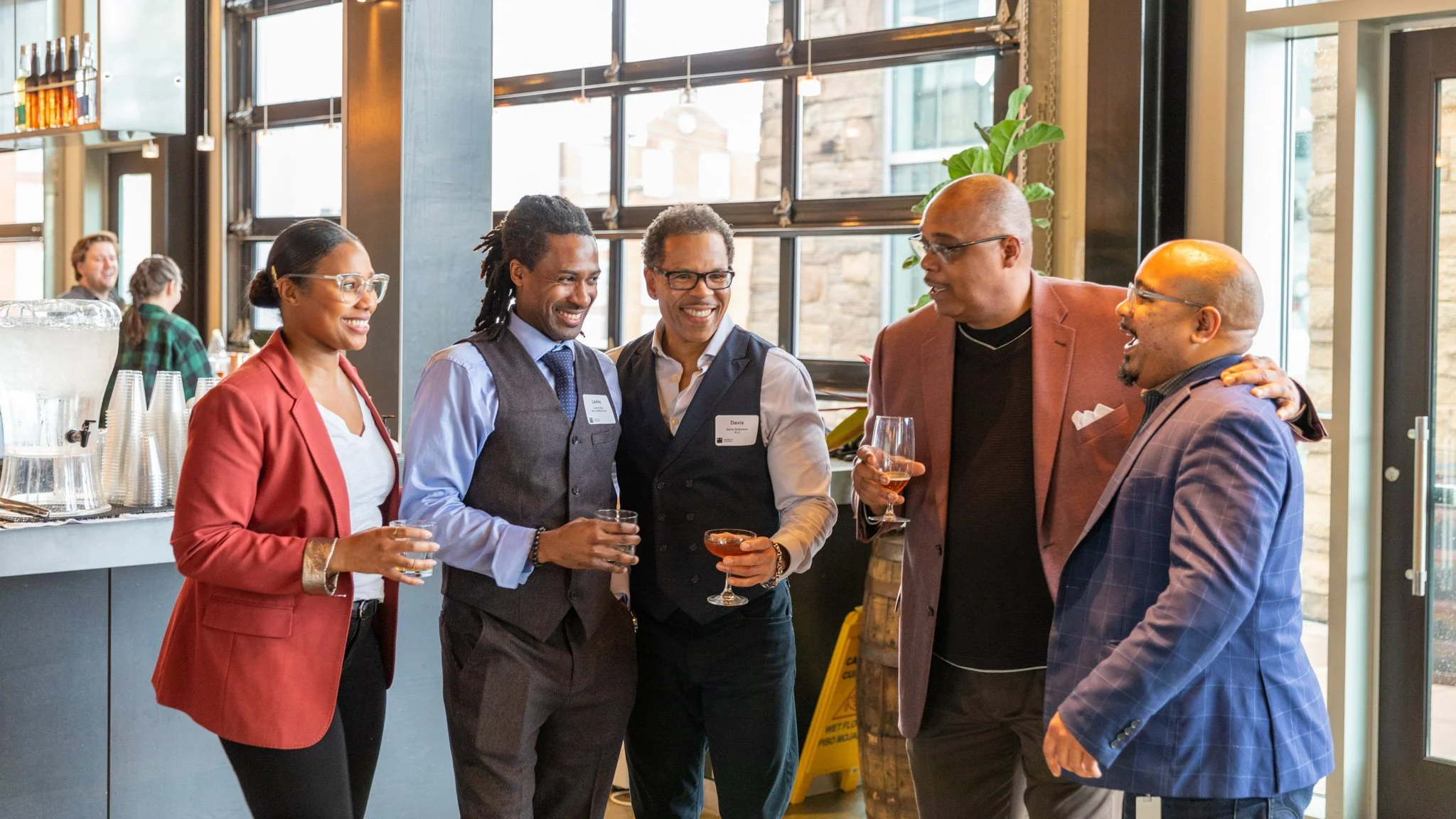 A group of five diverse professionals smiling and talking at a social gathering in a modern indoor setting, with large windows, drinks in hand, and a bar area in the background.