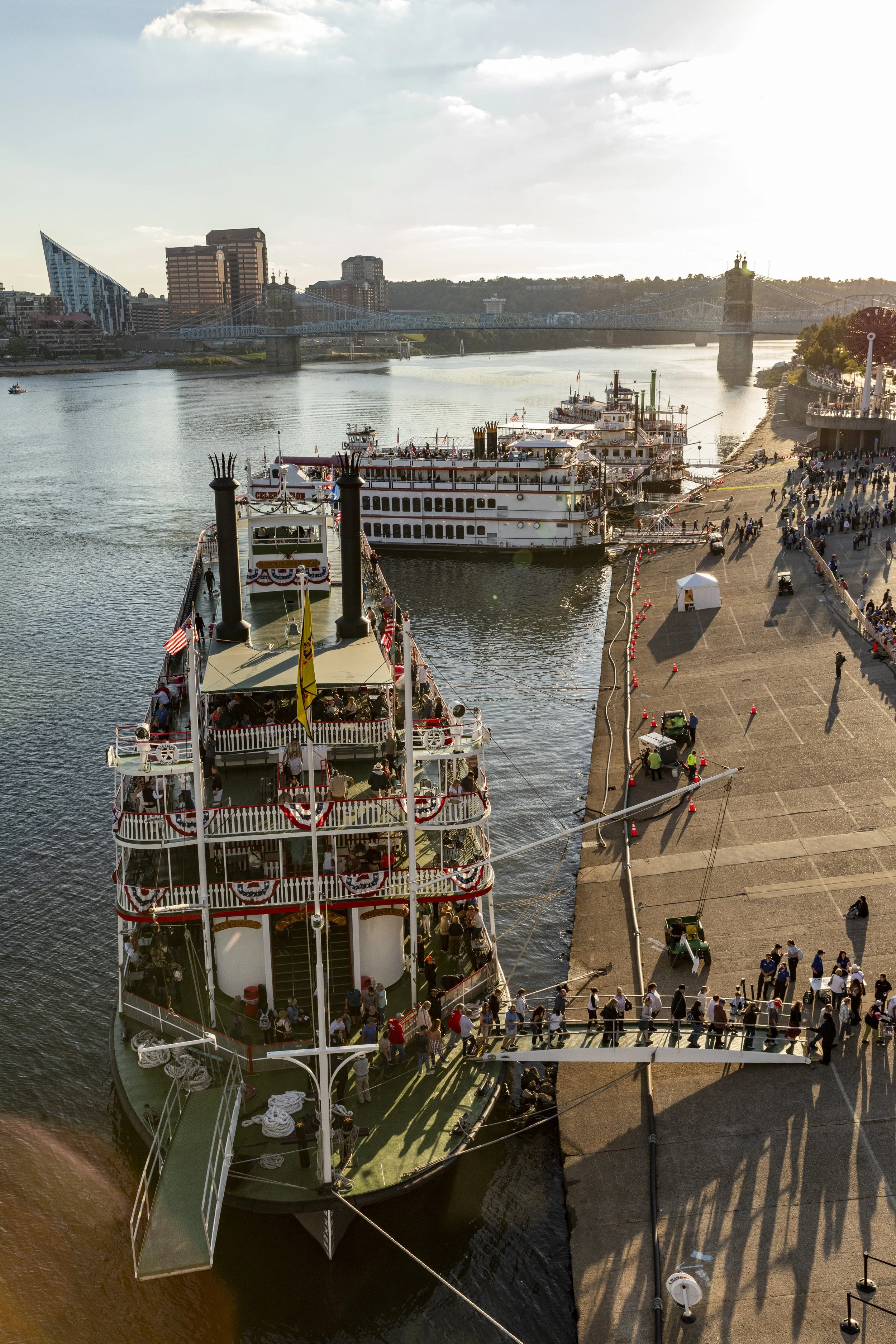 A river dock with several parked boats and a crowd of people walking along the pier in a cityscape, with a bridge and modern buildings in the background during sunset.