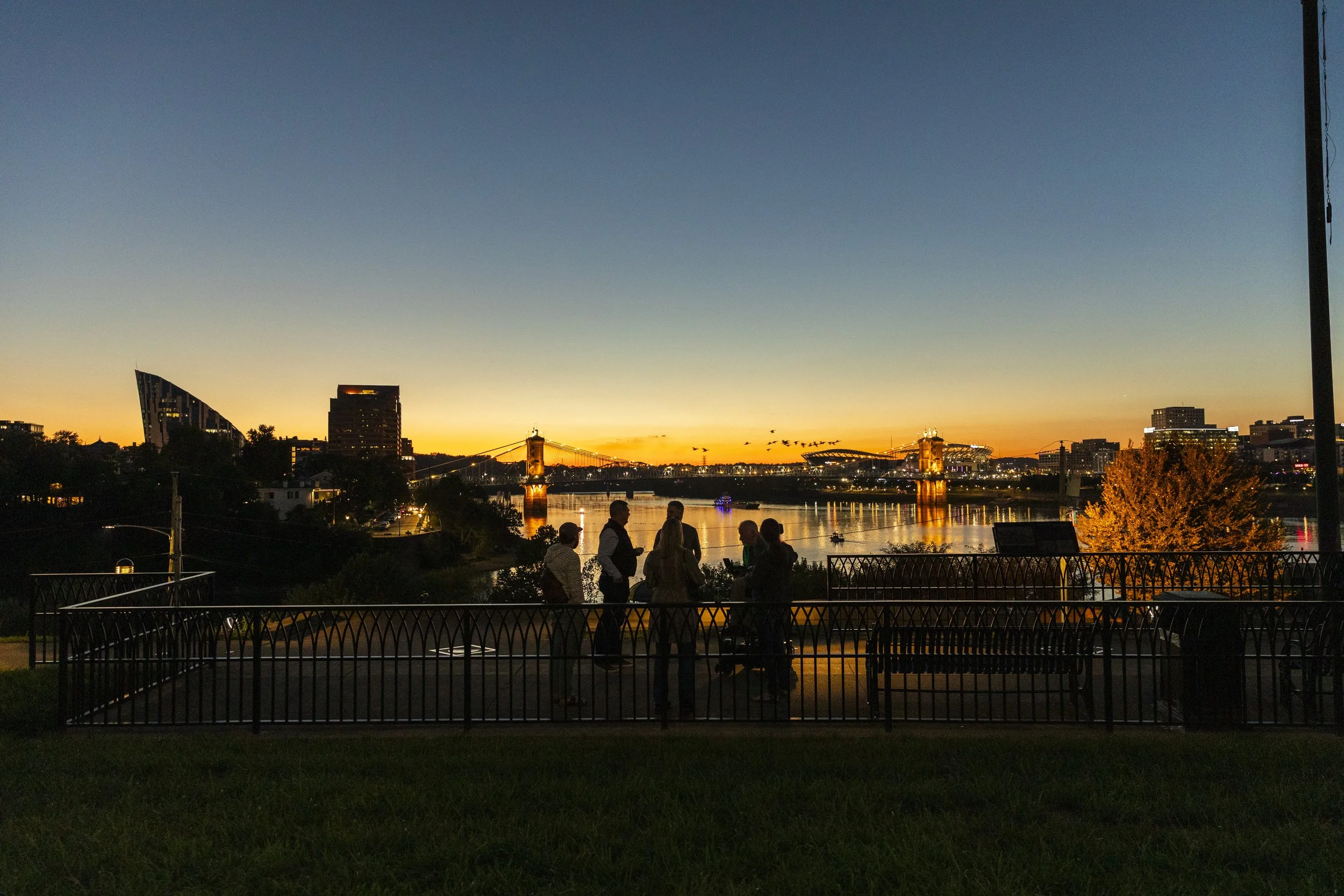 Silhouettes of people chatting on a riverside promenade at sunset with bridges and city skyline in the background.