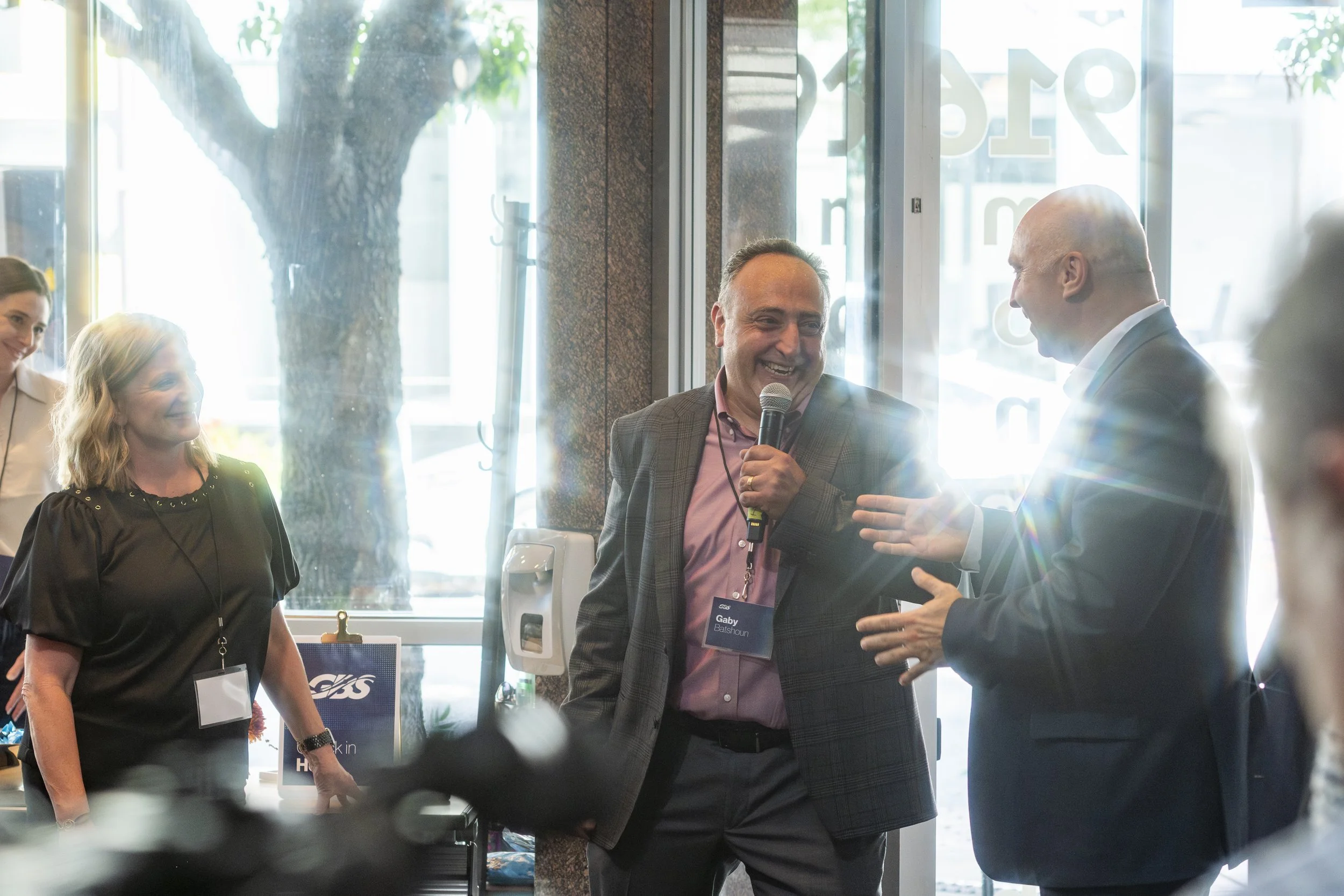 People at a business event, two men are engaged in conversation, with one holding a microphone, inside a bright venue with glass doors and a large tree visible outside.