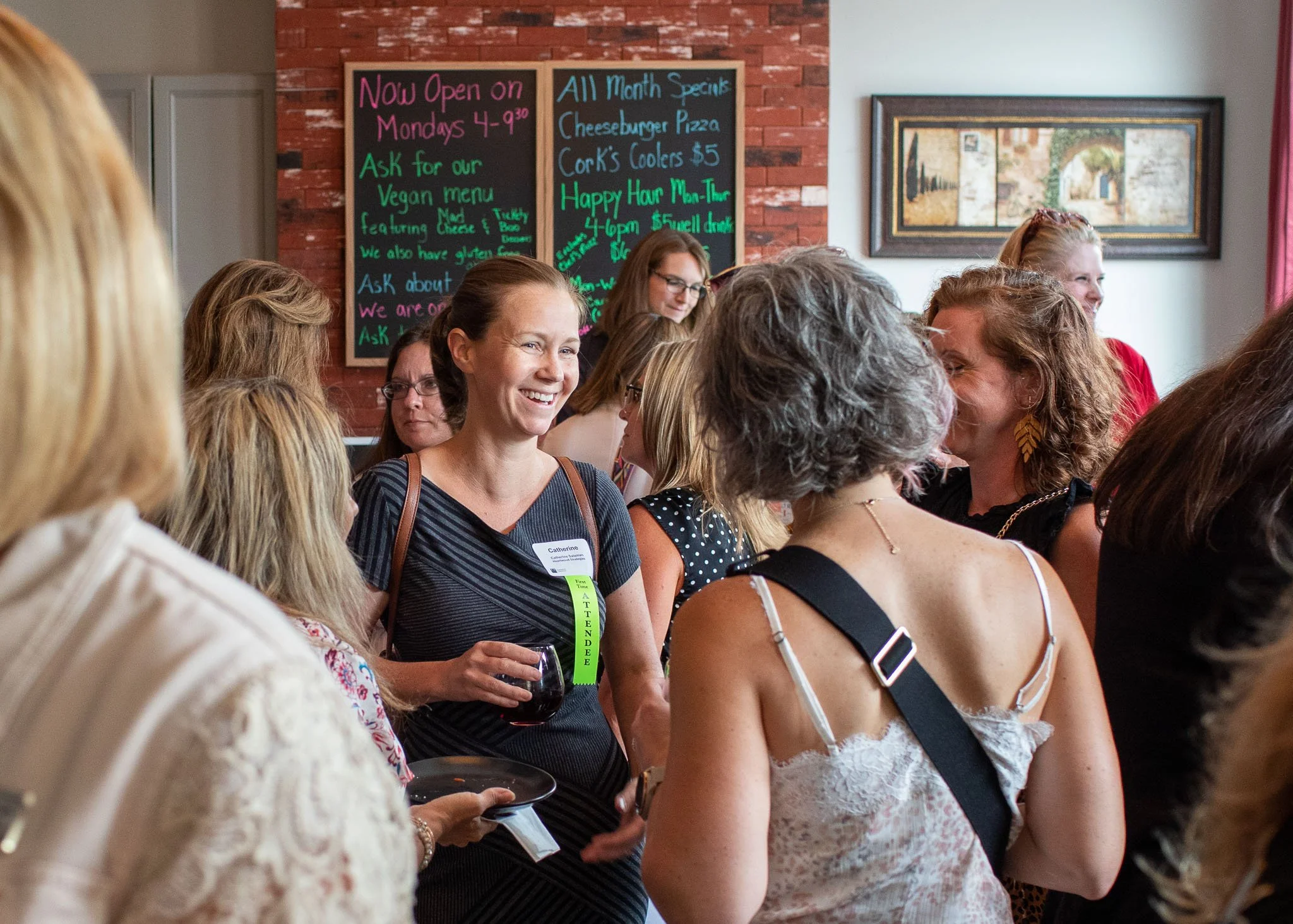 A group of women gathering and socializing indoors, with a woman holding a glass of red wine smiling and talking.