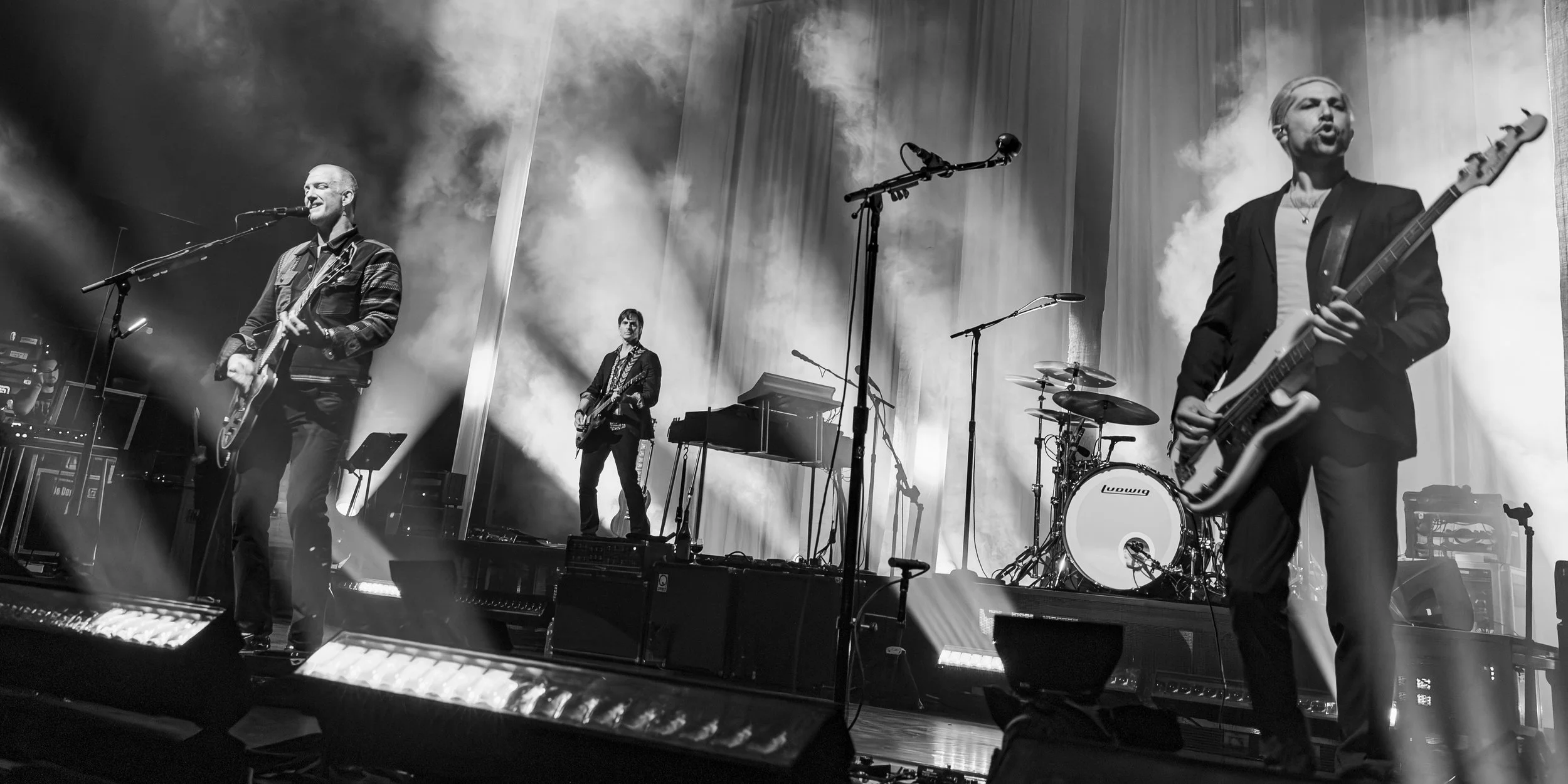 A four-member band performing on stage, with fog and bright lights. Two guitarists in suits are in the front, one with a bass guitar and the other with an electric guitar, singing into microphones. A keyboardist and a drummer are in the background.
