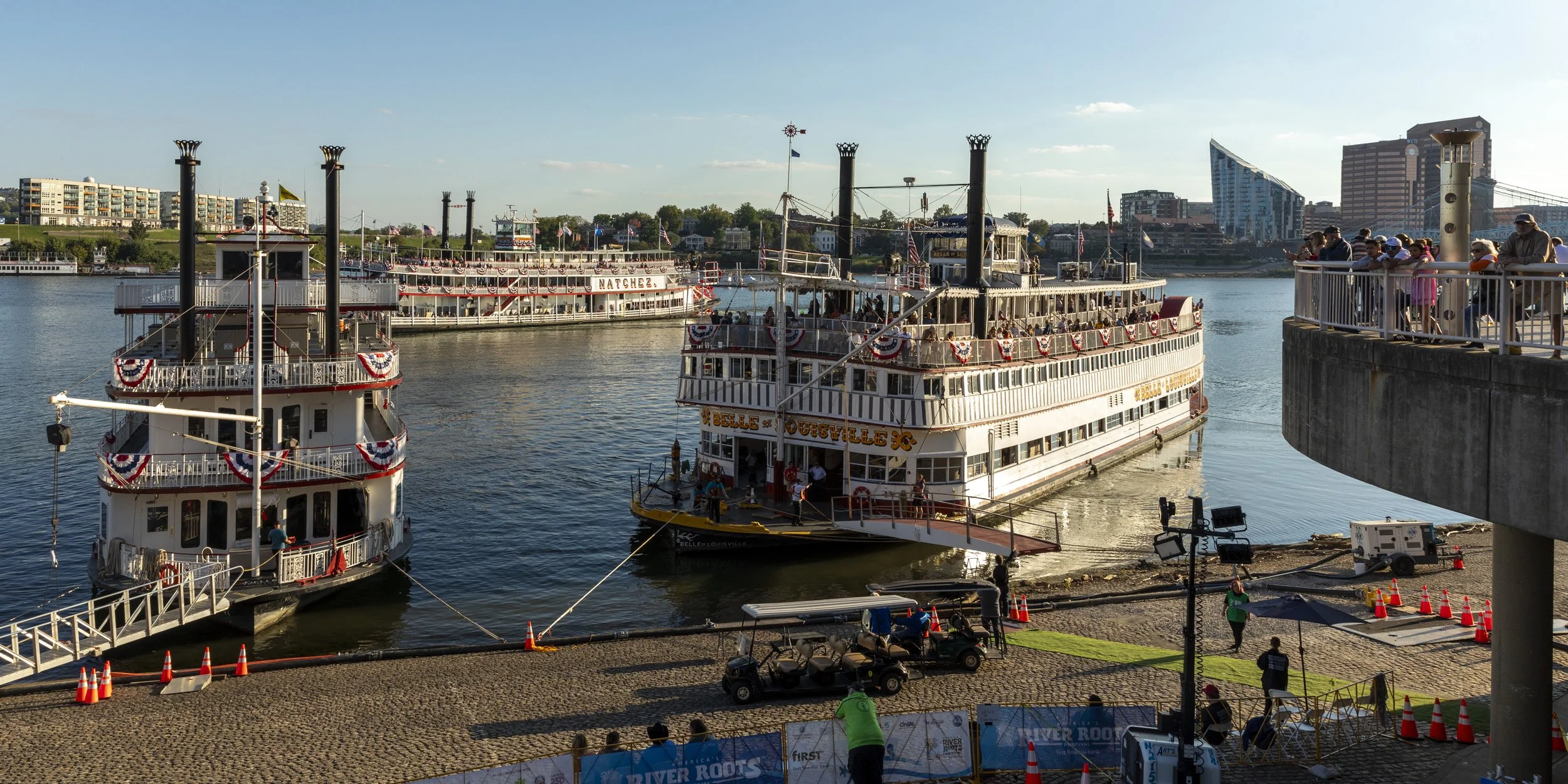 Three historical paddle wheel riverboats docked at a pier on a river, decorated with red, white, and blue bunting, with tourists on board and on the pier, overlooking city buildings under a clear blue sky.