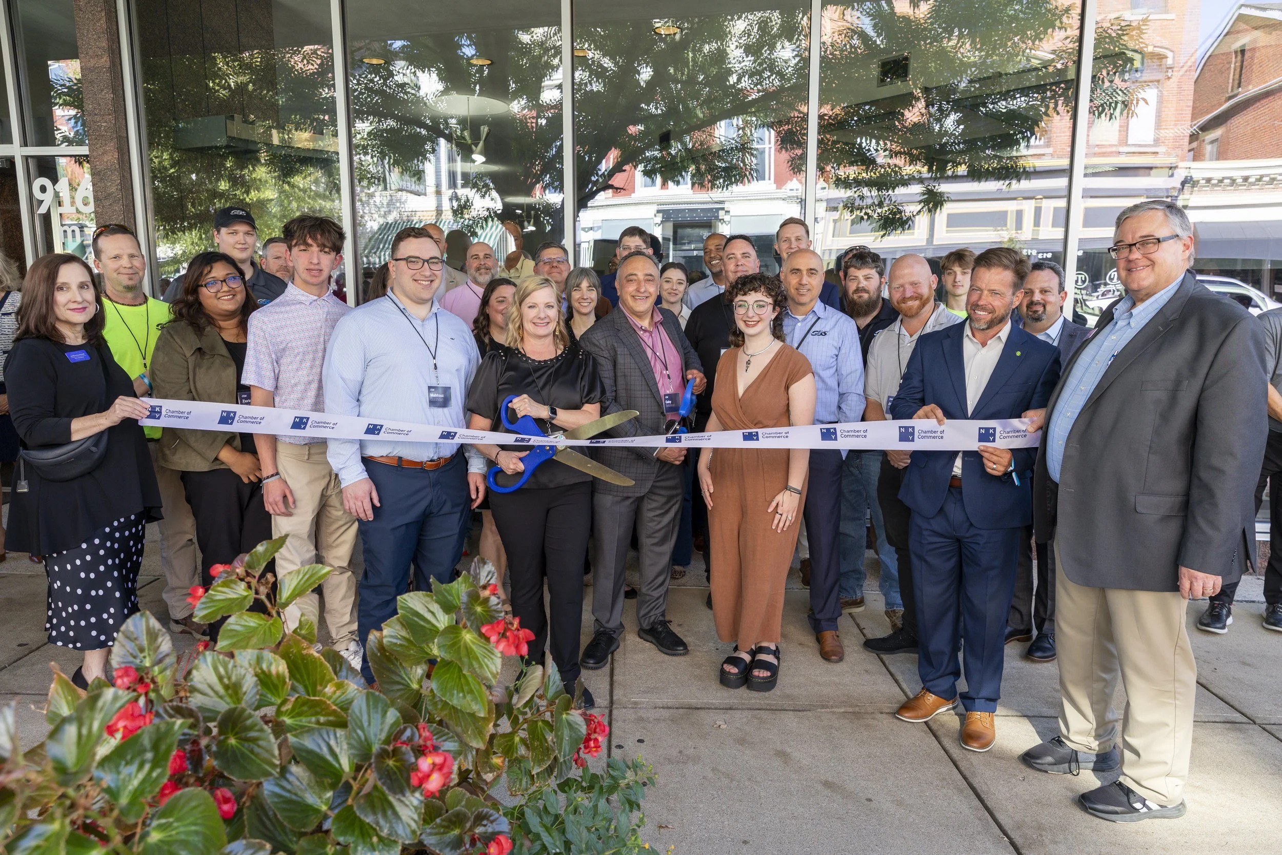 Group of people participating in a ribbon-cutting ceremony outside a building, with some holding scissors, celebrating a grand opening or special event.