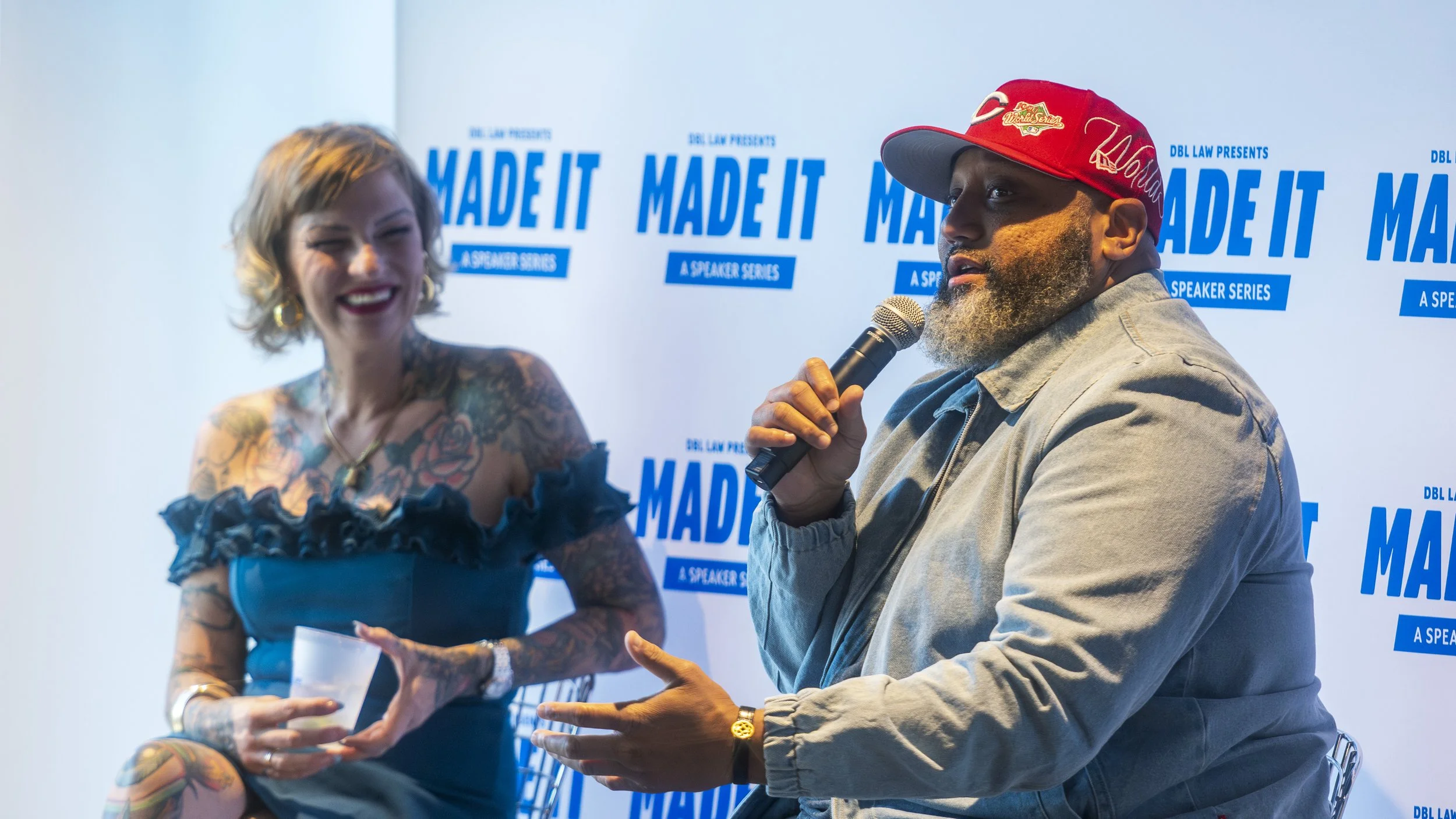 Two people seated, a woman smiling, holding a cup, and a man speaking into a microphone, with a backdrop that reads 'Made It' and 'A Speaker Series'.