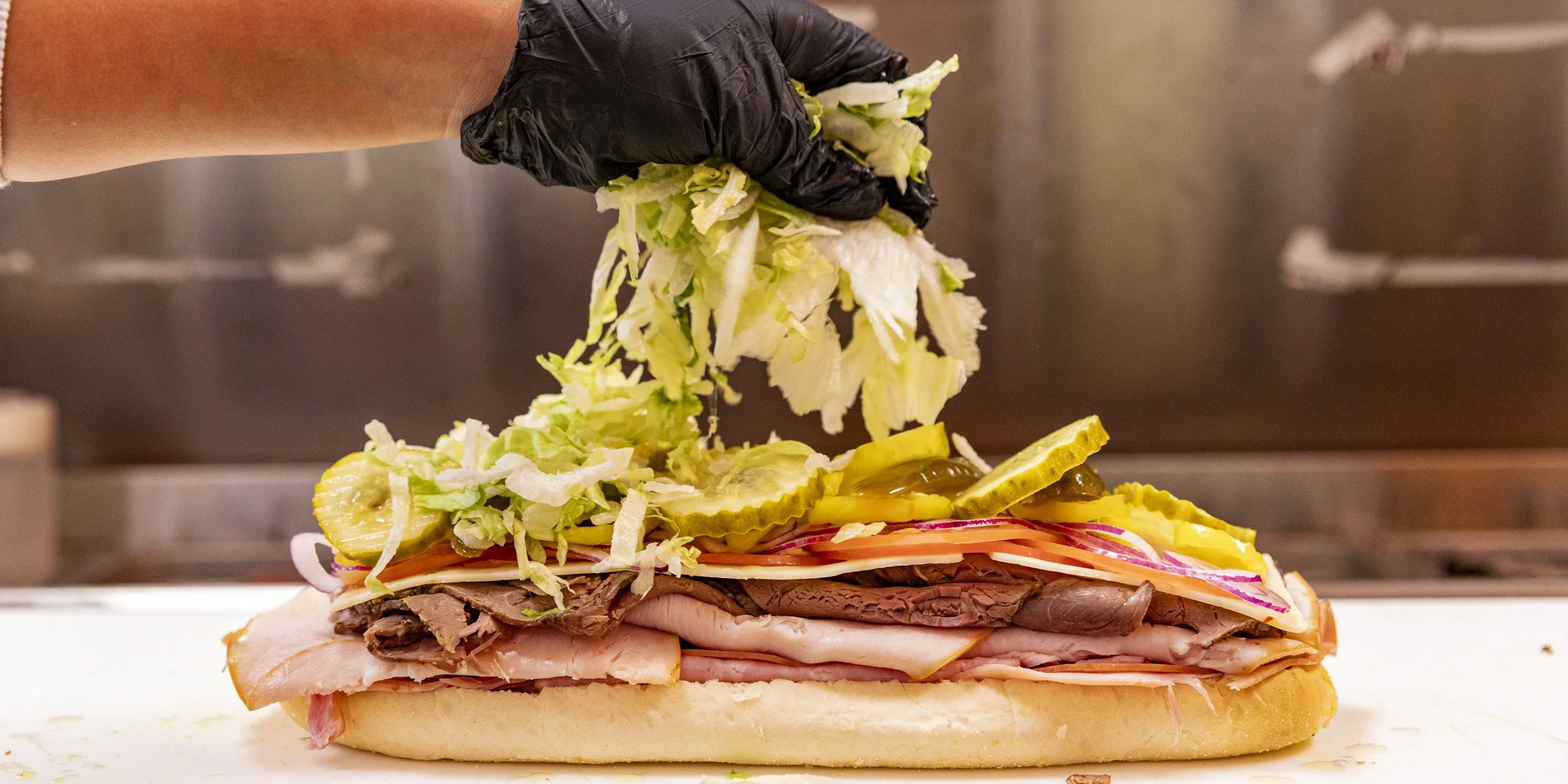 A person wearing a black glove adding shredded lettuce and other vegetables onto a large sandwich or sub with various meats, pickles, and vegetables on a bun.