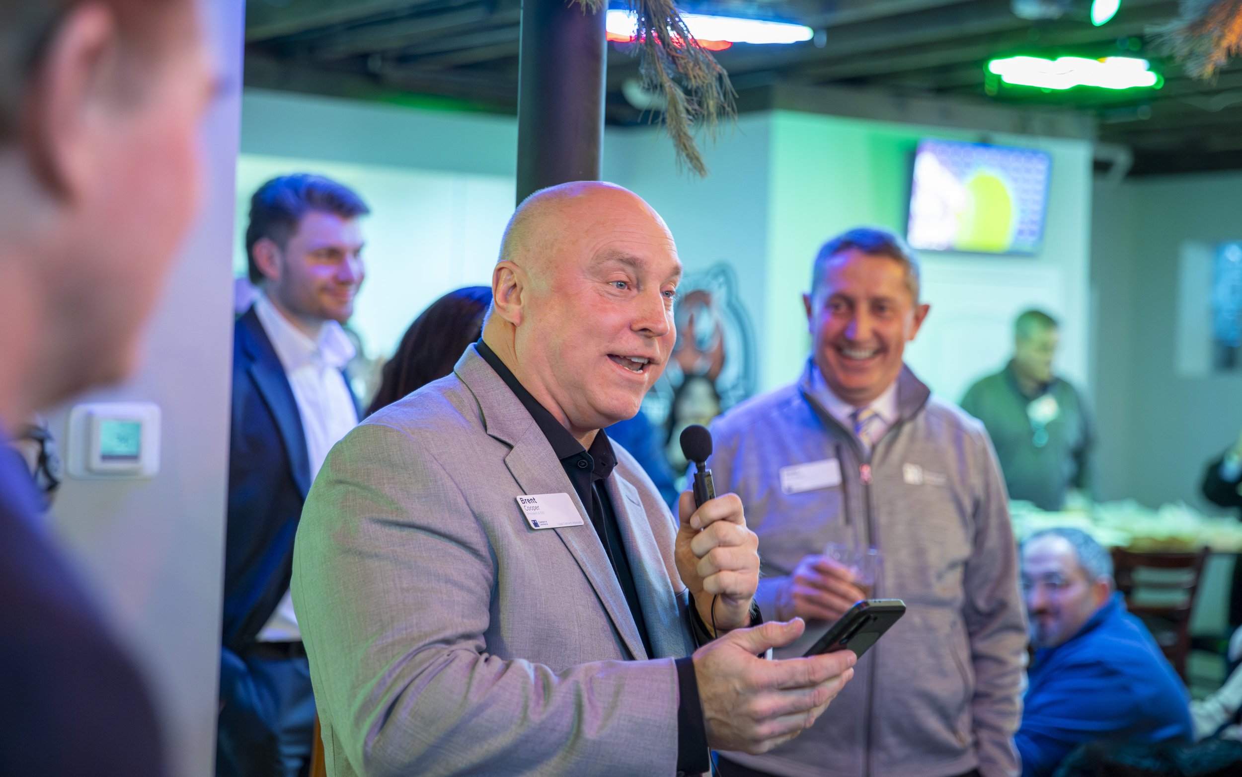 A man in a gray blazer holding a microphone and a smartphone, speaking to a group of people in a lively indoor setting with neon lighting.