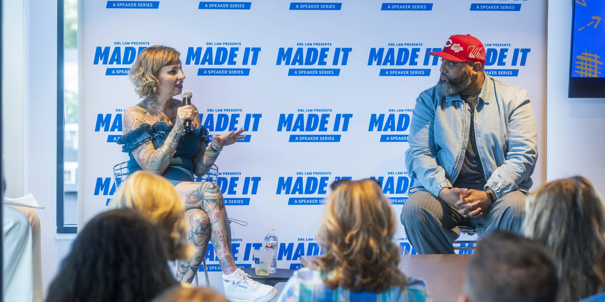 A woman with tattoos sitting with a microphone talking to a man in a red cap at a speaker series event. Behind them is a blue and white backdrop with the words 'MADE IT' and 'A SPEAKER SERIES' repeated.