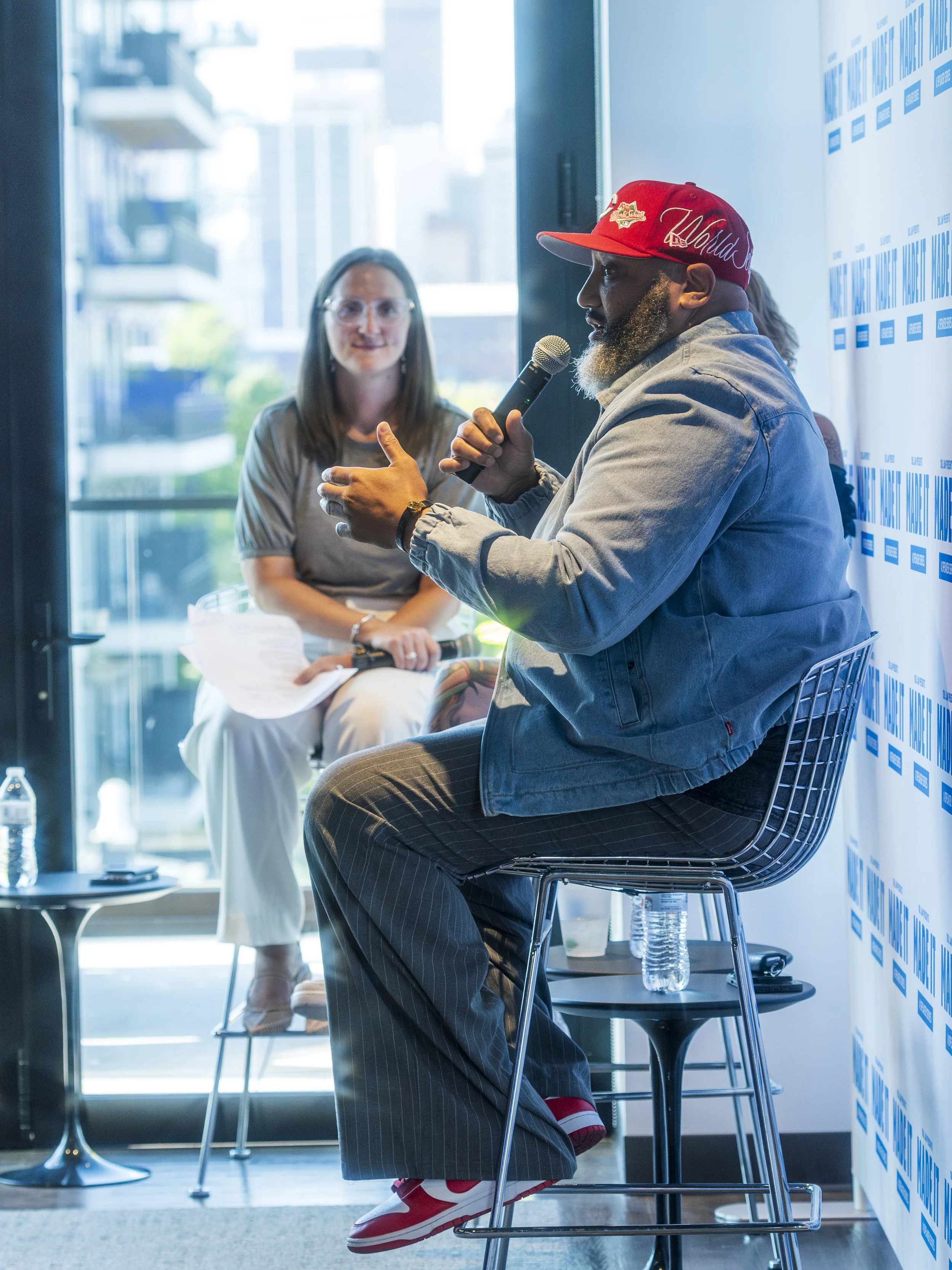 A man with a red cap and denim jacket speaking into a microphone during a panel discussion. A woman is sitting in the background, holding papers and listening.