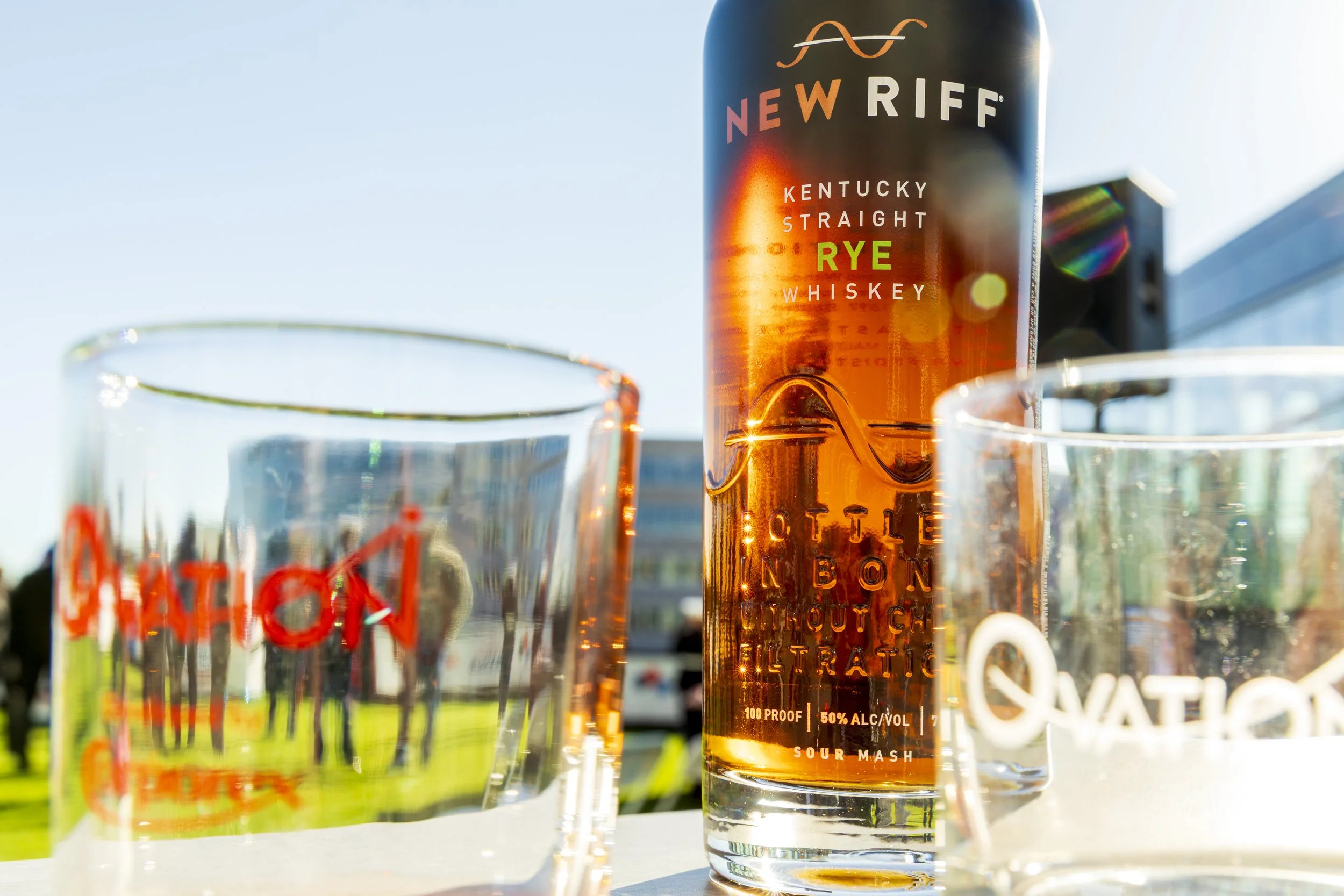 A can of New Riff Kentucky Straight Rye Whiskey positioned between two glasses, with a grassy outdoor background and sunlight reflecting off the glass surfaces.