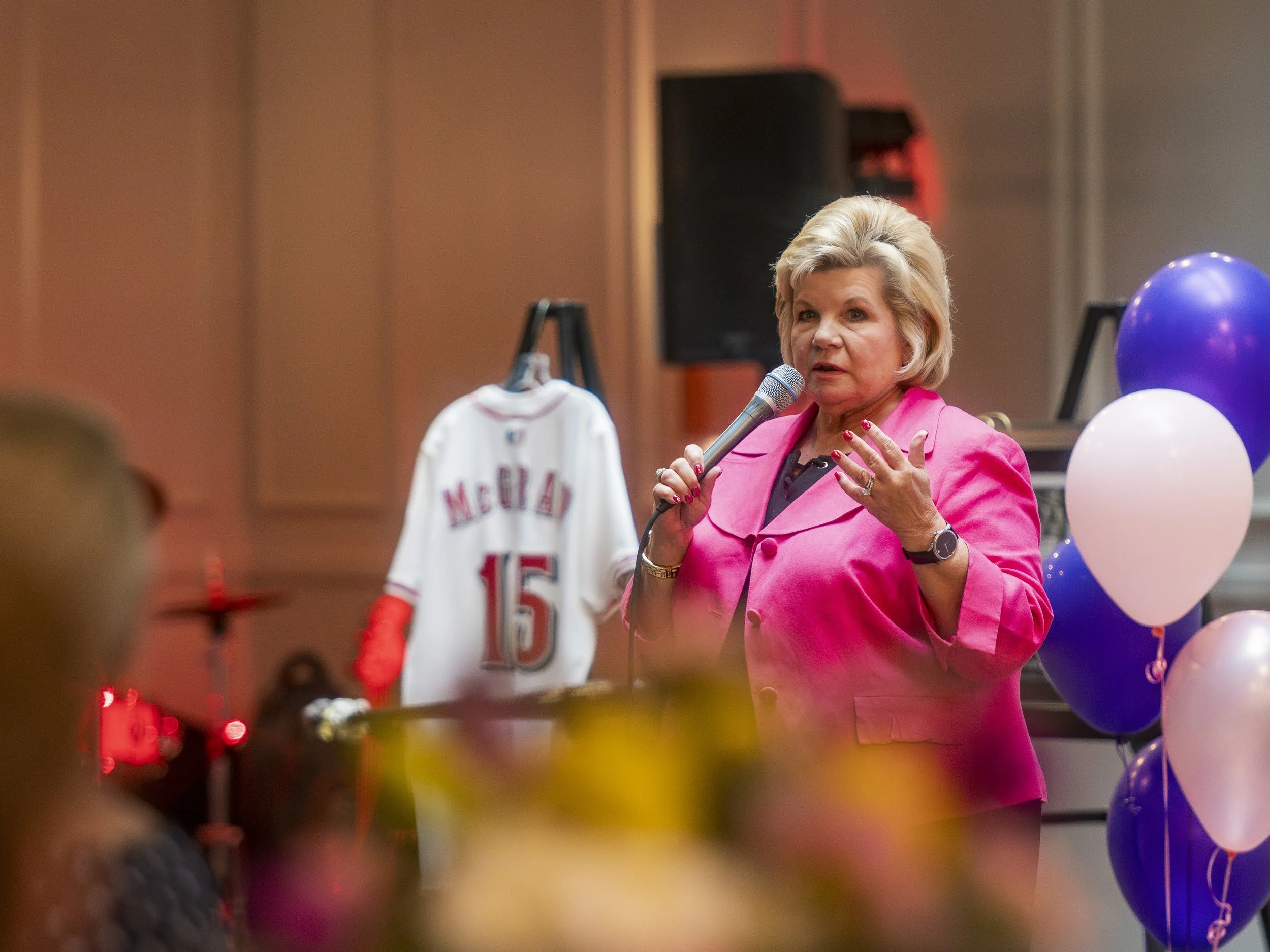 A woman with blonde hair wearing a pink blazer speaking into a microphone at a rally or celebration event, with balloons and a hockey jersey hanging in the background.