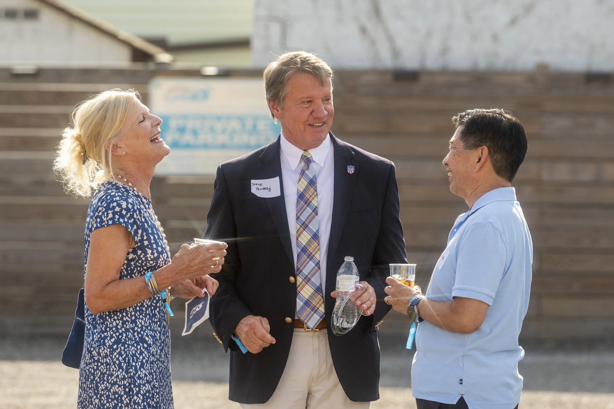 Three people, two men and one woman, are standing outdoors, engaged in conversation and smiling. The woman is wearing a blue patterned dress, the man in the middle is wearing a dark blazer and tie with a name tag, and the other man is in a light blue
