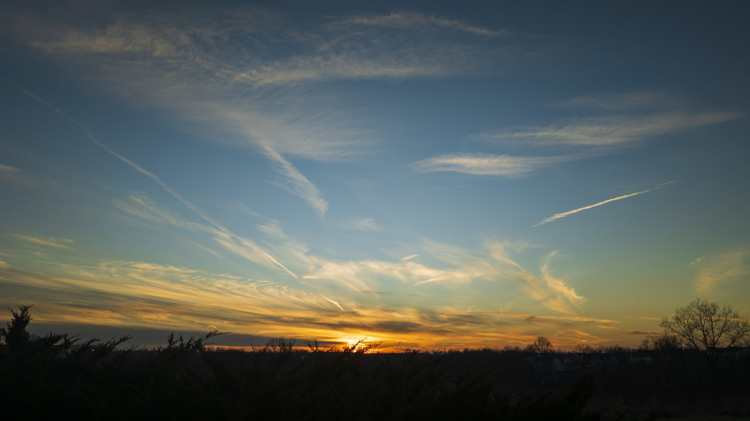 Sunset over a landscape with a dark silhouette of trees and bushes in the foreground, and a sky with scattered clouds and contrails.