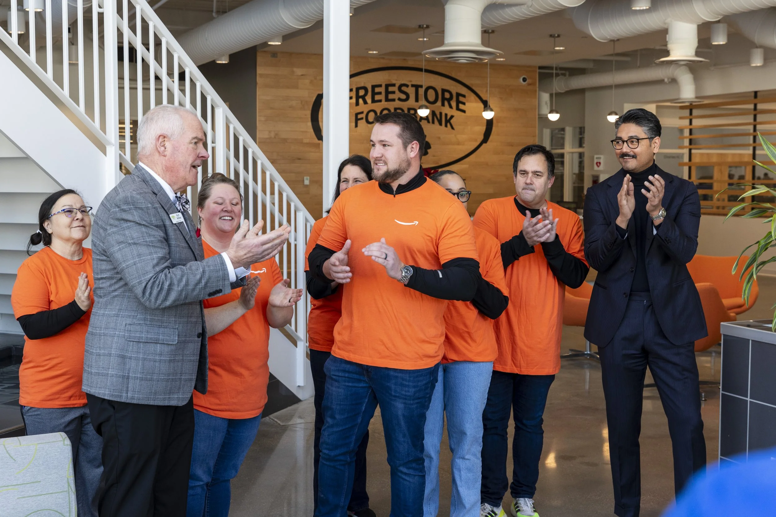 Group of people at a food bank, some wearing orange shirts, standing inside a modern building with a staircase and a sign that says 'Freestore Foodbank', and clapping.