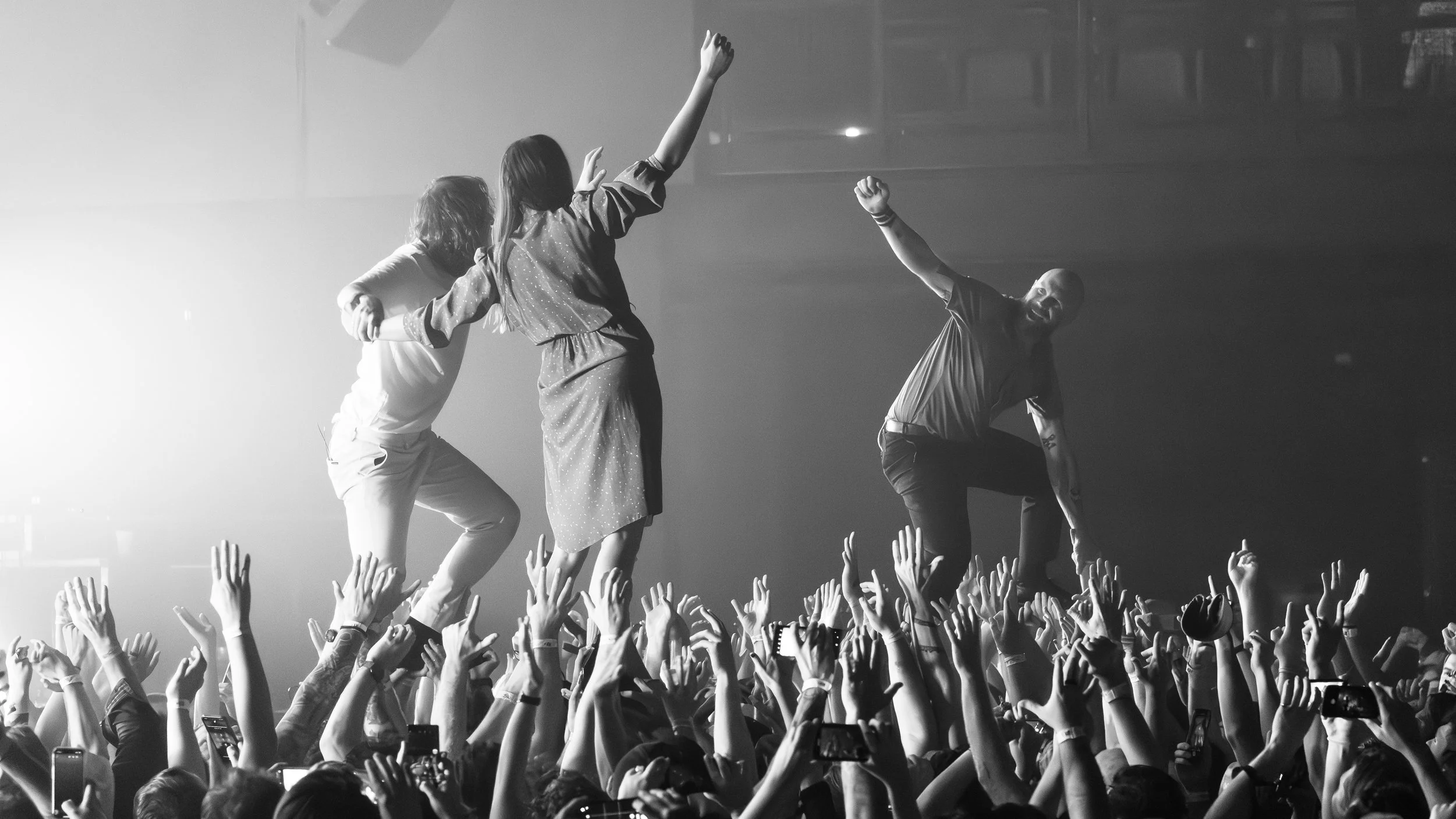 Black and white photo of three people on a stage, engaging with an audience, with many audience members holding up their hands and taking photos.
