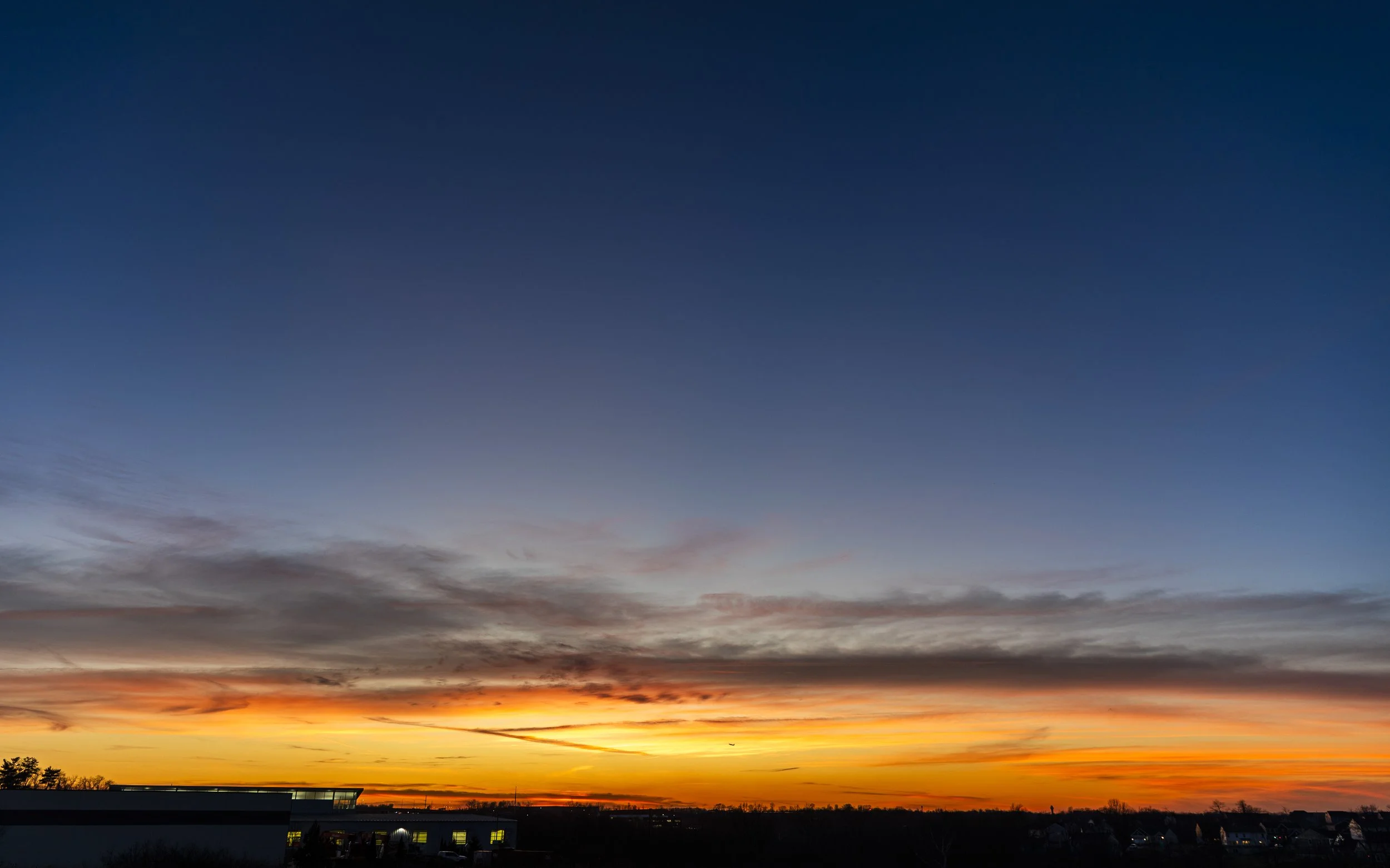 Sunset over a horizon with orange, pink, and yellow colors, dark silhouettes of buildings and trees, and a partly cloudy sky with shades of blue and gray.