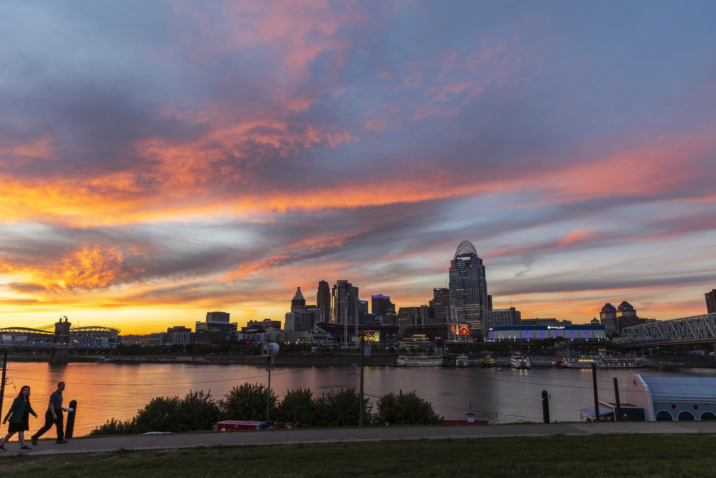 Sunset over Cincinnati skyline with colorful sky, river in foreground, and people walking on a park path.