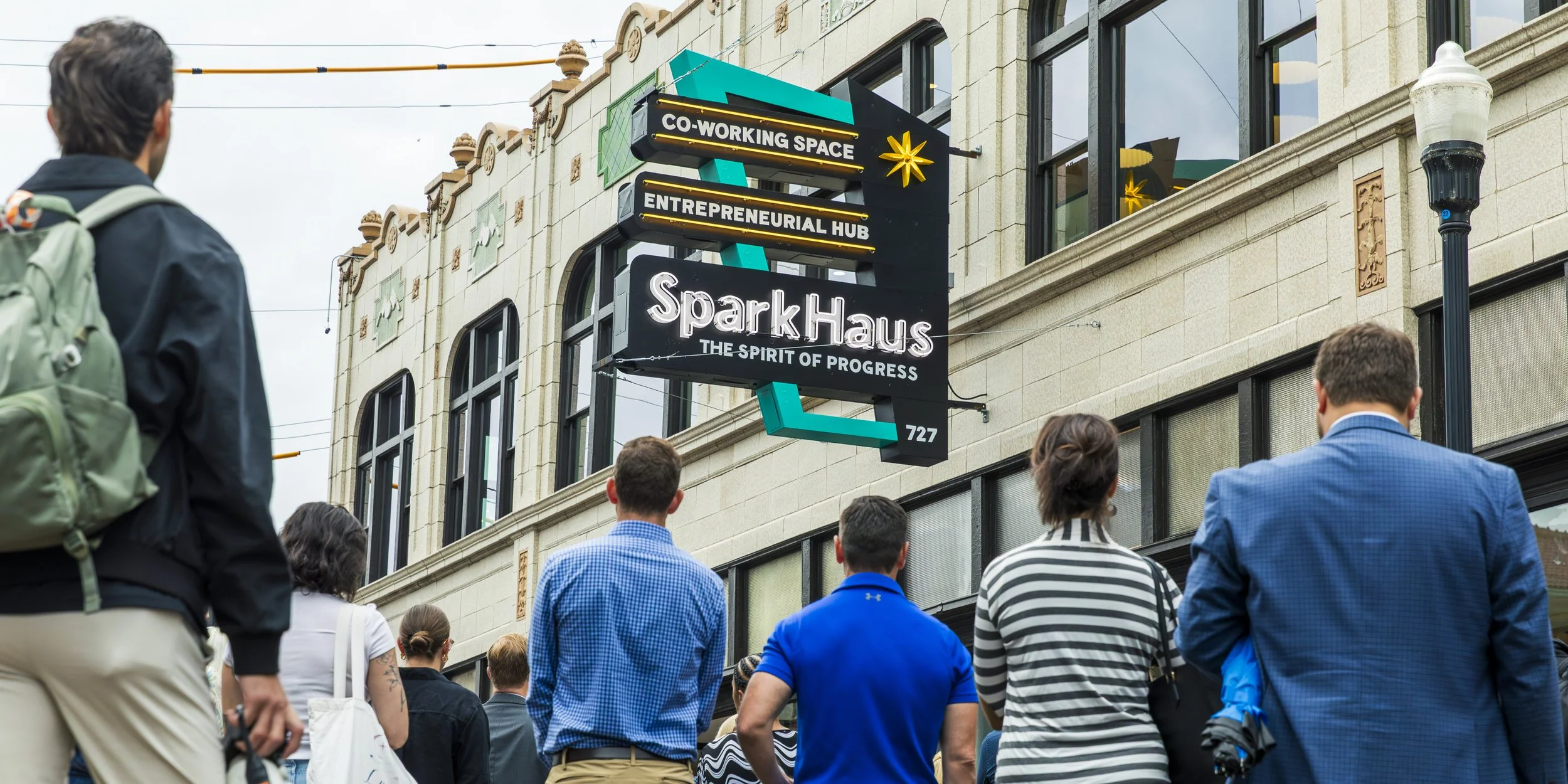 People standing on a city sidewalk looking up at a sign for Spark Haus, a coworking space and entrepreneurial hub, on an ornate building with large windows. The sign reads 'CO-WORKING SPACE,' 'ENTREPRENEURIAL HUB,' and 'THE SPIRIT OF PROGRESS.'
