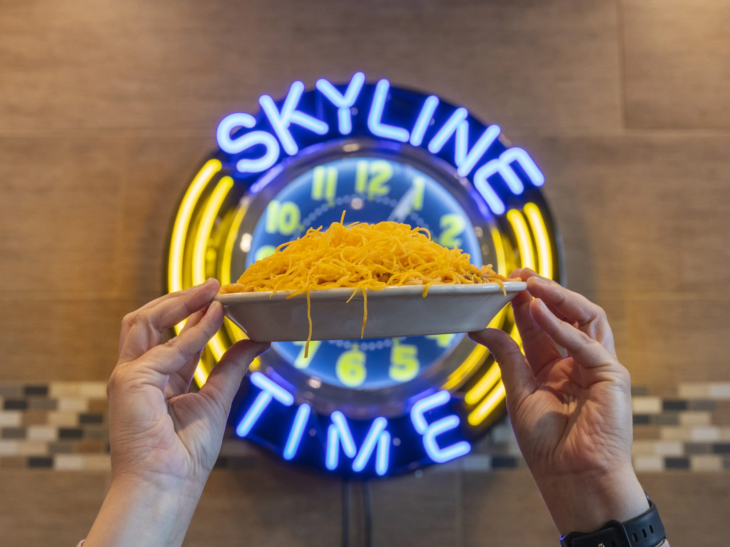 Hands holding a white tray of cheese-covered spaghetti pasta with a neon sign that says 'SKYLINE TIME' behind them.