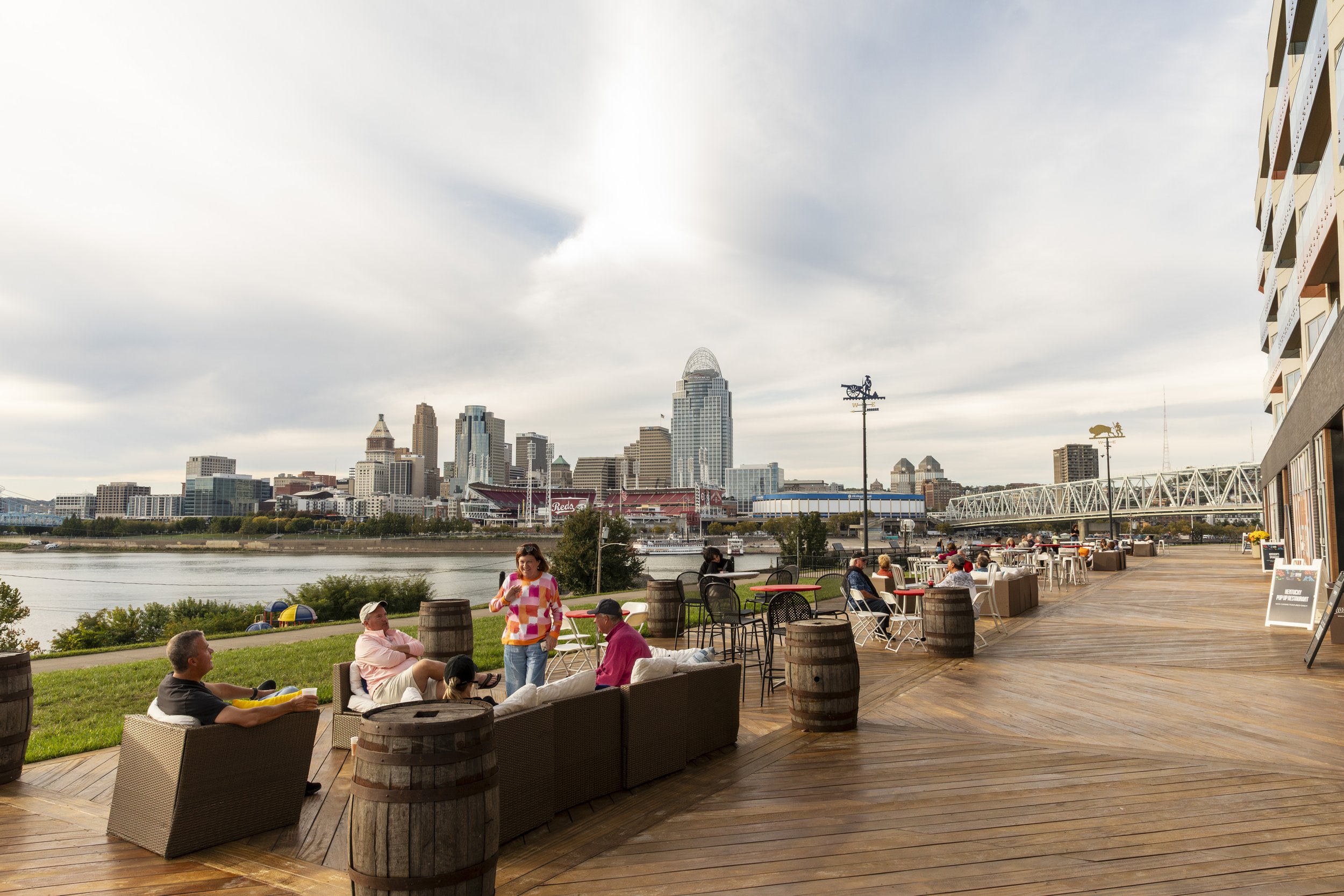 People relaxing and socializing on a wooden outdoor patio with city skyline in the background, across a river.