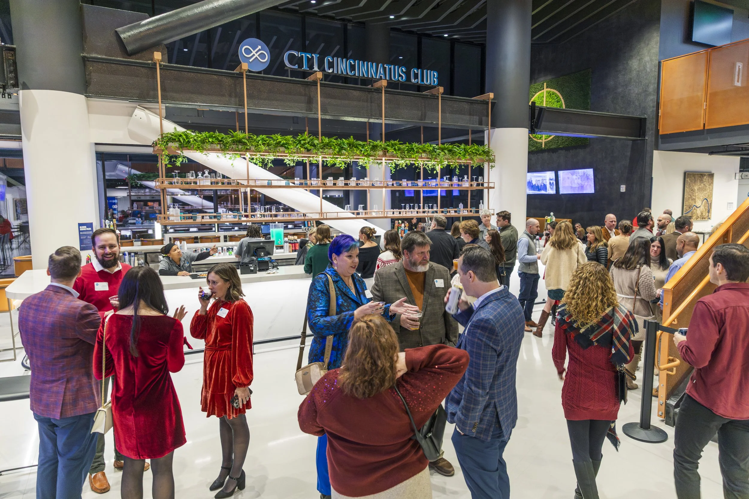 A group of people attending a social event at a modern indoor venue with a bar, where they are talking and drinking. The background features a sign that says 'CTI Cincinnati's Club' and a contemporary, stylish interior design.