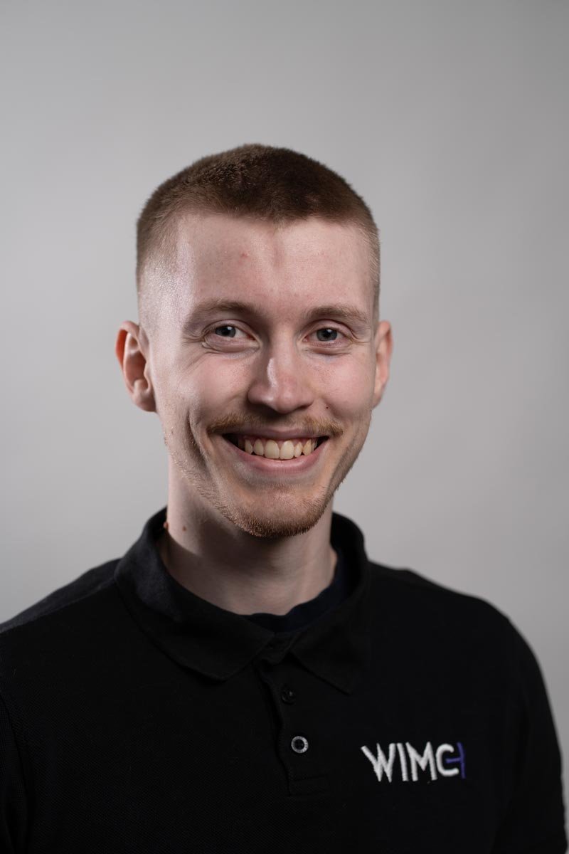 Ung man med kort, ljusbrunt hår, leende och gröna ögon, iklädd svart polo med WIMG på bröstet, grå bakgrund.