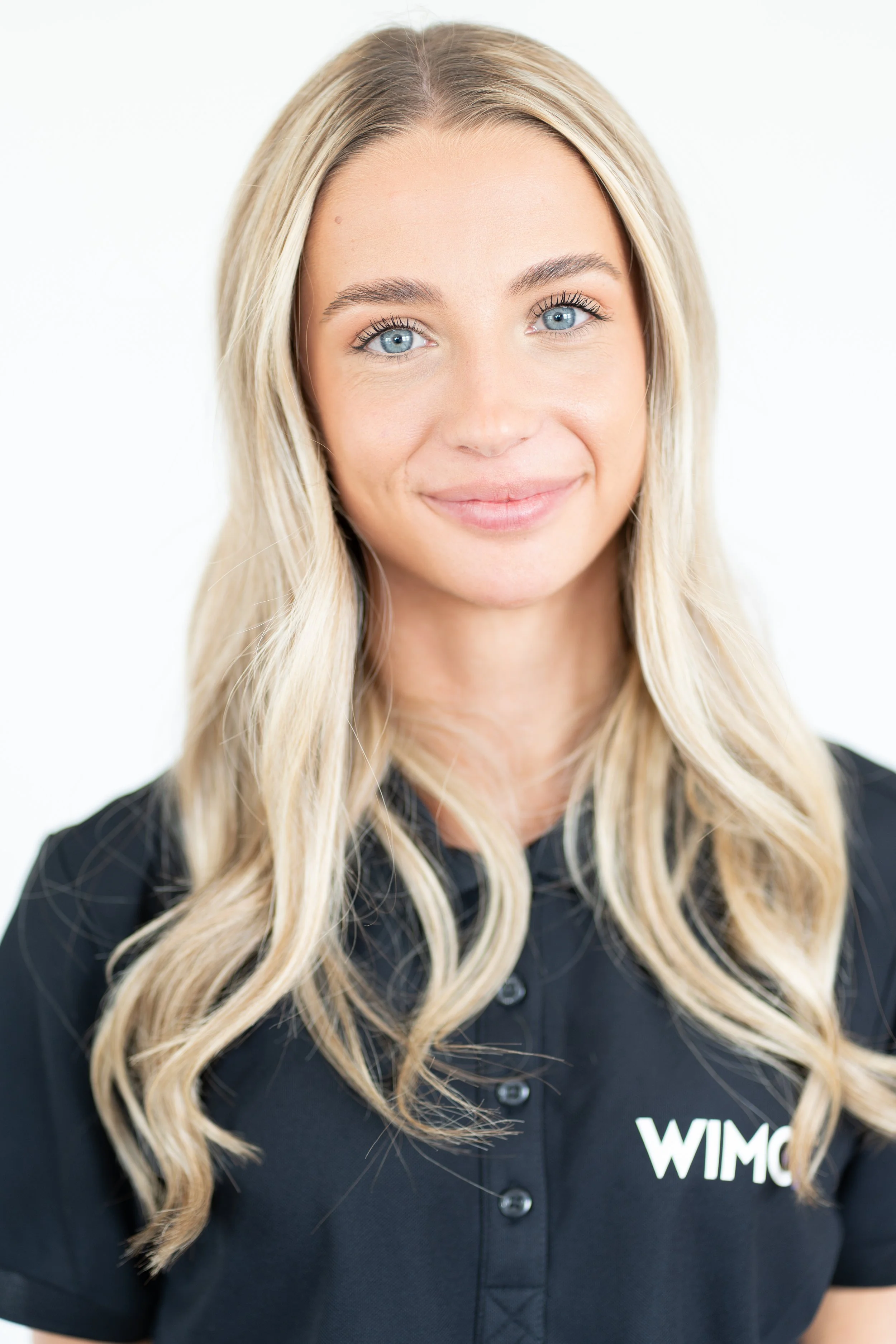 Ung kvinna med långt blont hår, blå ögon och svart WIMC-amarbjëdsplagg som ler mot kameran mot en vit bakgrund.