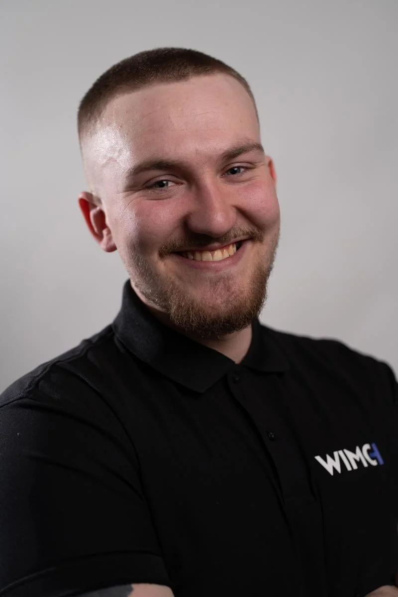 Ung man med kort hår, skägg och leende, bär svart LAN-tröja med WIMC-logo mot en ljus bakgrund.