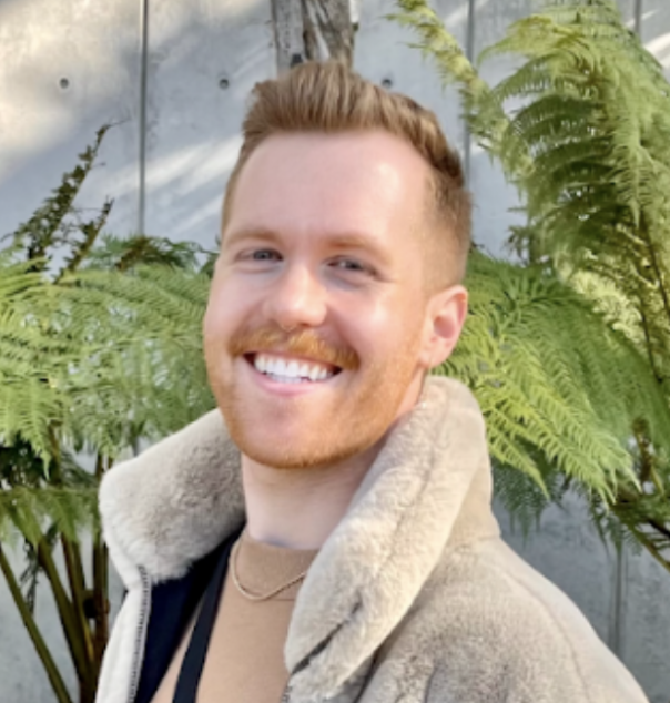 A smiling man with short, reddish hair and a beard stands outdoors in front of large green ferns and a gray wall.