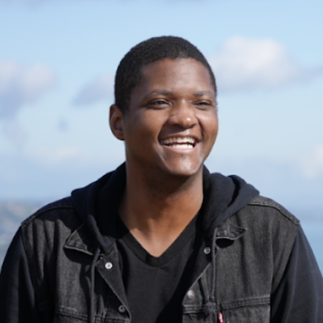 A young man with short black hair smiling outdoors against a cloudy blue sky background.