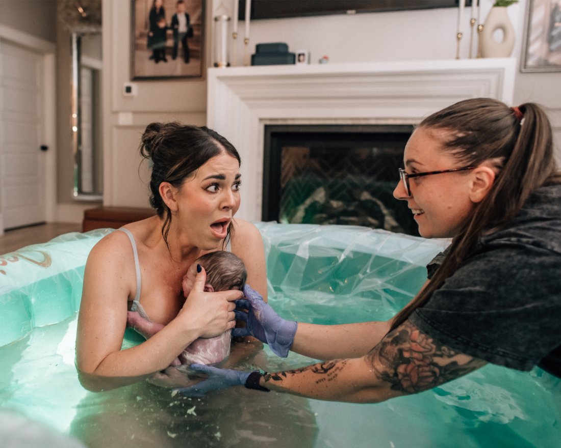 Birthing parent holding their newborn in a birth pool while doula provides reassurance, capturing the emotional moments that often inspire becoming a doula.