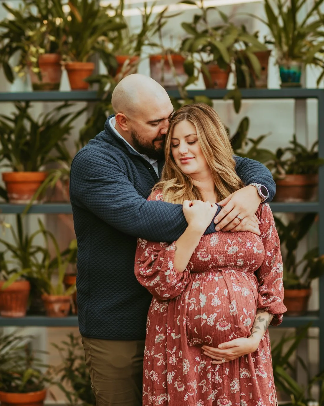 Expecting couple standing together in a greenhouse, sharing a quiet embrace as the pregnant mother rests her hand on her belly during maternity photography Fort Collins.
