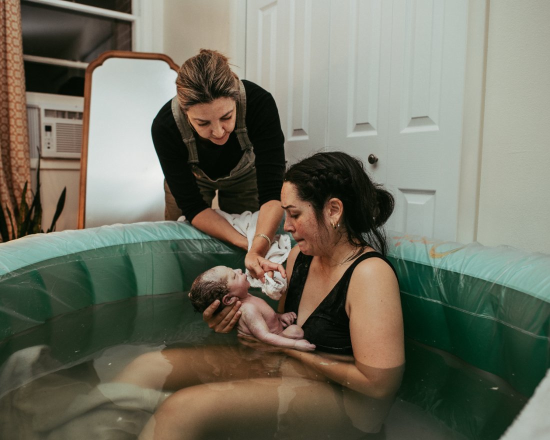Doula supporting a birthing parent as they hold their newborn in a birth pool, showing hands on care and presence central to becoming a doula.