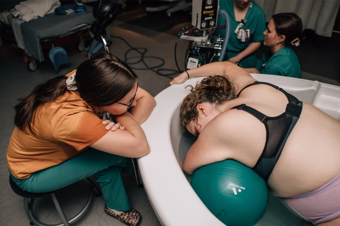 Doula sitting close and offering steady presence to a laboring parent resting over a birth tub, showing the quiet support involved in becoming a doula.