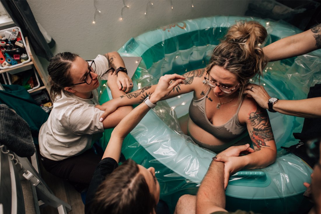 Birthing parent laboring in a birth pool surrounded by support people and hands on comfort, illustrating support preferences outlined in a birth plan checklist.