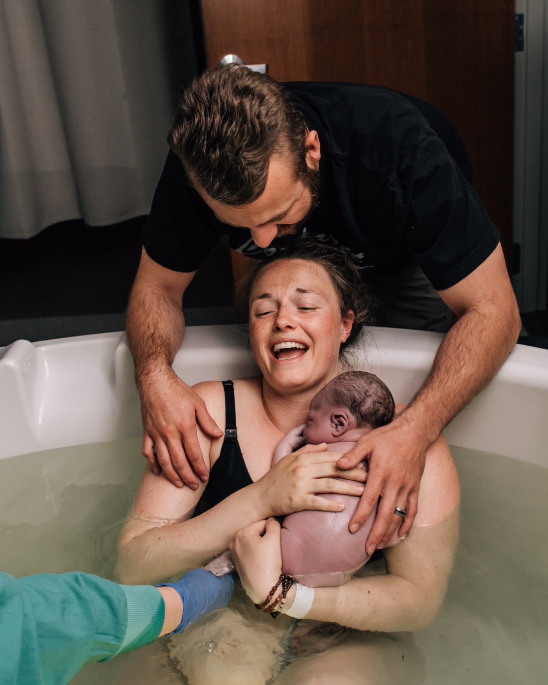 Birthing parent smiling while holding their newborn in a birth tub with partner support nearby, photographed through childbirth photography that documents emotional connection.