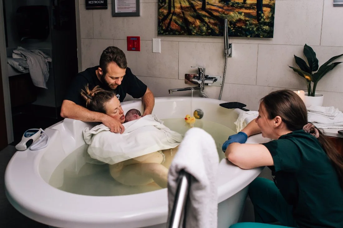 Mother in a birth tub holding her newborn while partner and Trauma Informed Doula look on in amazement