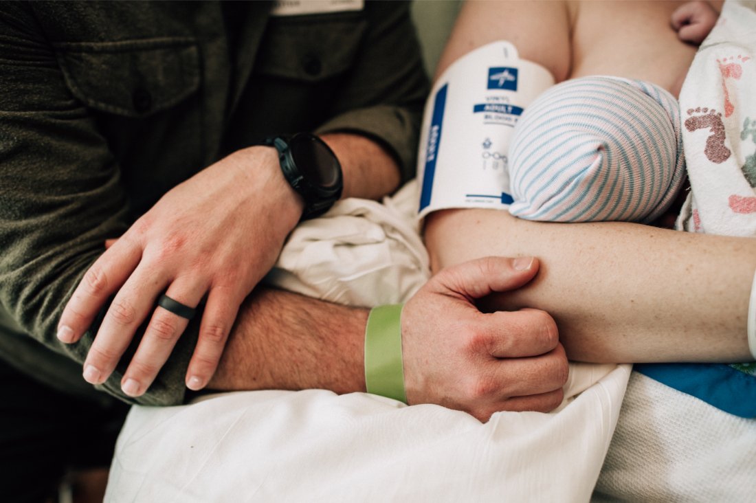 Parent holding newborn in a hospital bed with partner’s arm nearby, highlighting postpartum and bonding preferences often noted in a birth plan checklist.