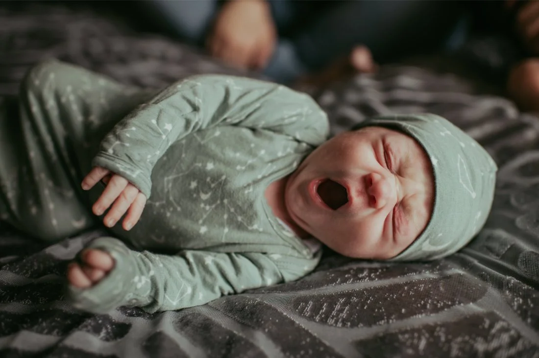 Newborn lying on a bed in a simple outfit mid stretch, documenting real moments through natural newborn photography.