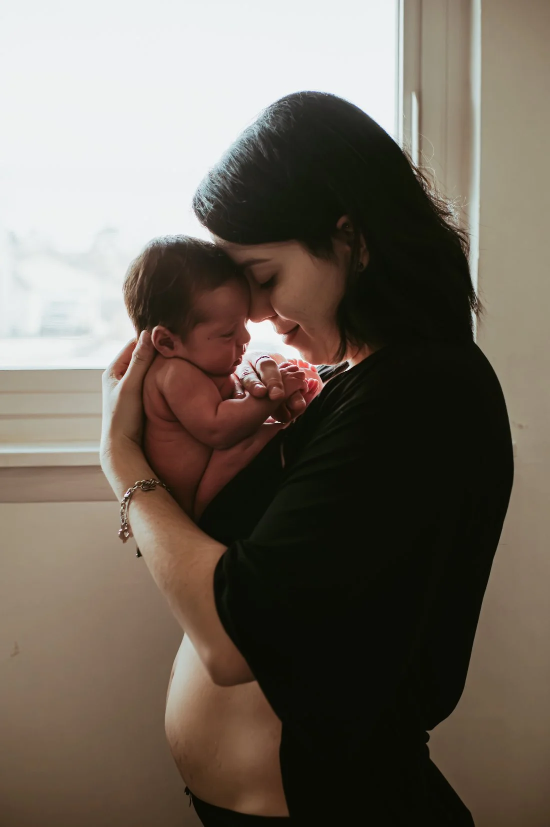 New mom and baby posing together in a softly lit window after birth with Trauma Informed Doula support nearby