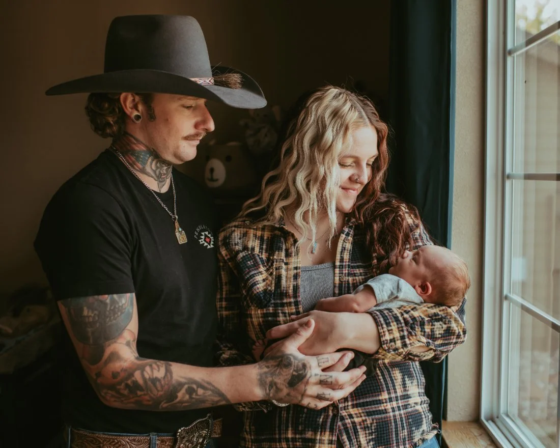 Parents standing together by a window holding their newborn, captured in a relaxed and unposed natural newborn photography moment.
