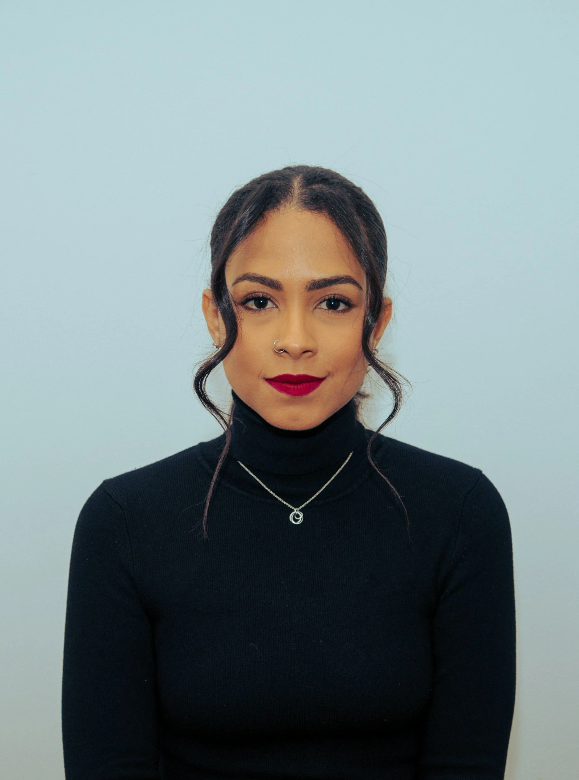 A young woman with dark hair styled in loose curls wearing a black turtleneck, red lipstick, and two necklaces, sitting against a plain light blue wall.