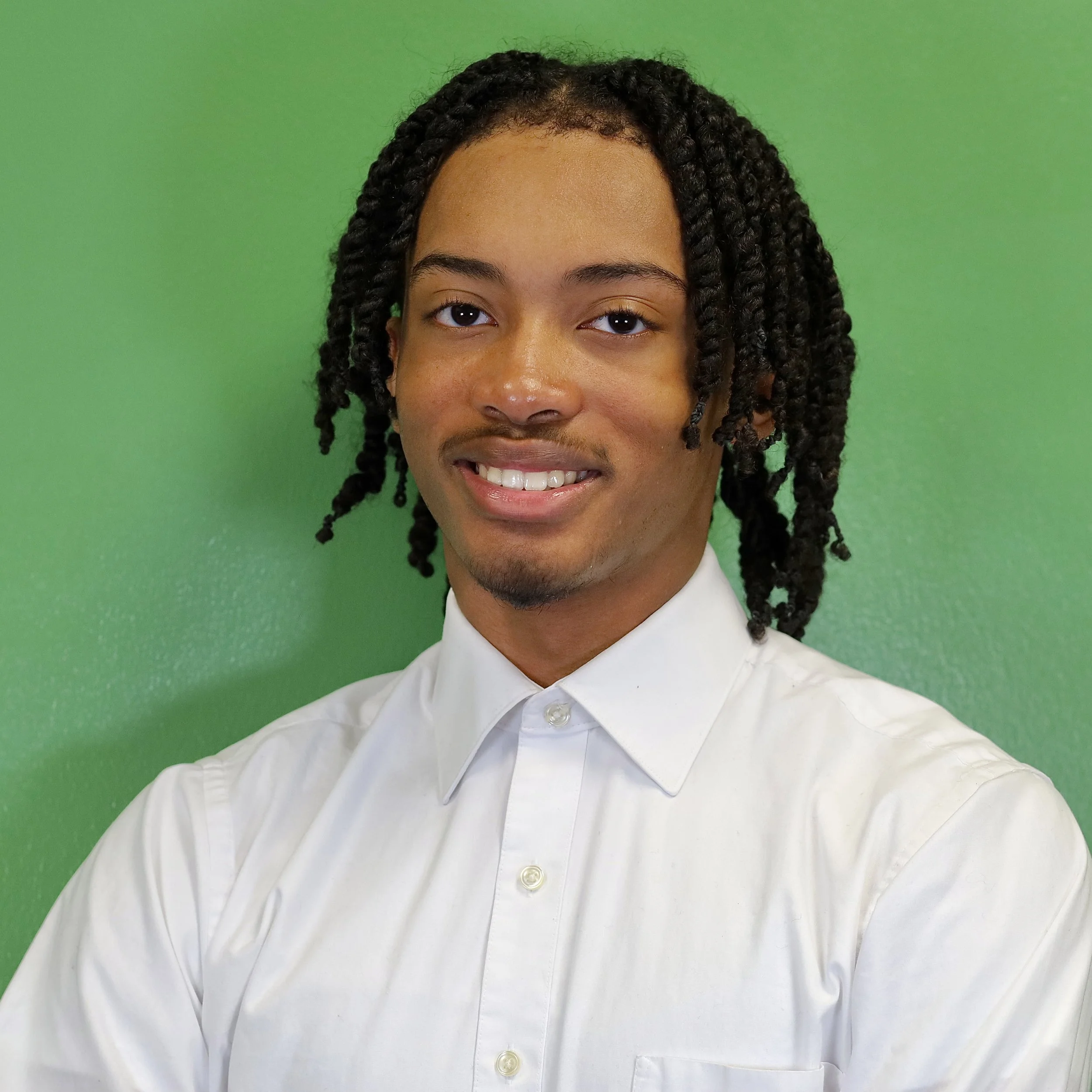 Portrait of a young man with dark curly hair, wearing a white button-up shirt, standing in front of a green background.