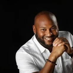 A smiling man with a beard wearing a white shirt, leaning forward with his hands clasped.