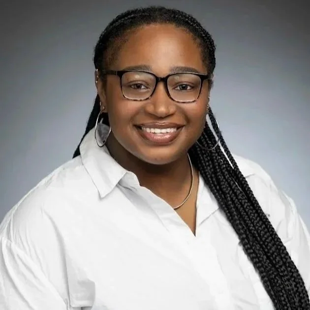 A young woman with dark braided hair, wearing glasses, hoop earrings, a white shirt, and a necklace, smiling against a gray background.