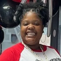 Smiling woman with curly hair and a red and white shirt, standing in front of gym equipment.