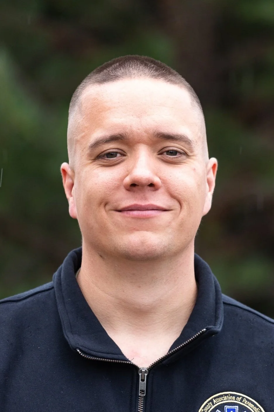 A person with short hair wearing a black jacket standing outdoors.
