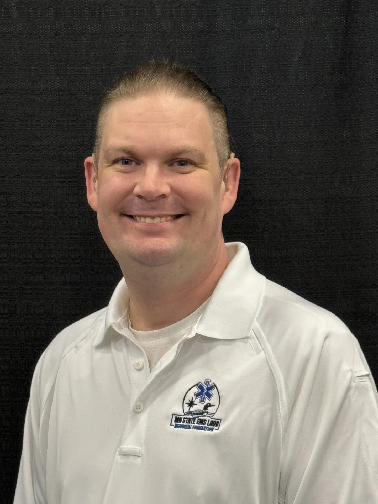 Smiling man in white polo shirt with logo against black background.